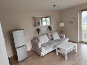 Ein Zimmer mit einem weißen Sofa, einem weißen Tisch, einem weißen Kühlschrank, einem Spiegel, einer Lampe und einem Fenster mit Jalousien.
