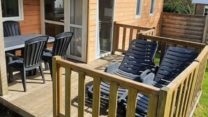 Eine Holzdar Terrasse mit Tisch und Stühlen, ein kleines Gebäude mit Schiebetür und ein eingezäunter Bereich mit Liegestühlen.