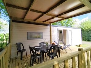 A covered outdoor area with a table and chairs, a small building with a sliding door, and a wooden deck railing. - Mobile home, pets allowed - 4 guests (Chaillé-les-Marais)