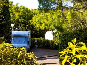 A white RV parked in a wooded area with dense trees and a gravel path. - Mobile home with AC - 6 guests (Lézignan-Corbières)