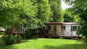 A mobile home with a porch and a fenced deck, surrounded by lush greenery. - Mobile home with AC - 6 guests (Monplaisant)