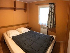 A single bed with a dark quilt, a bedside table, a wall-mounted shelf, a window with patterned curtains, and a radiator. - Mobile home, pets welcome - 4 guests (Perros-Guirec)