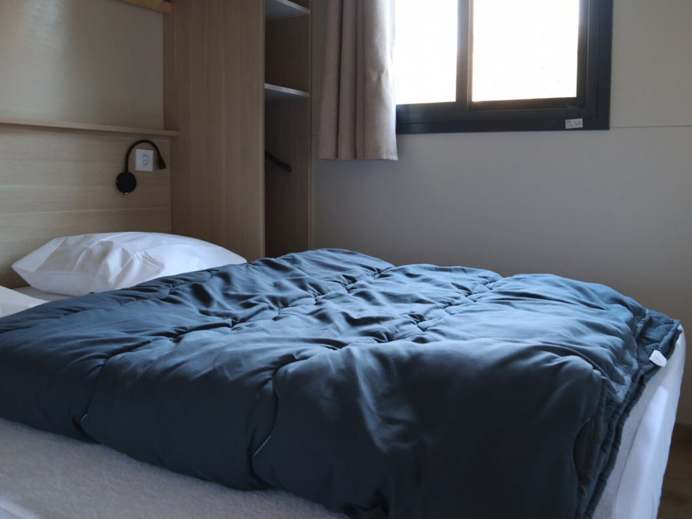 A bed with a blue duvet, a white pillow, a wooden headboard, a shelf with a black object, and a window with curtains.