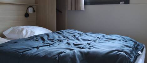 A bed with a blue duvet, a white pillow, a wooden headboard, a shelf with a black object, and a window with curtains.