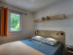 A bedroom with a bed, a window with curtains, a bedside table, and a shelf above the bed. - Mobile home, pets allowed - 10 guests (Groléjac)