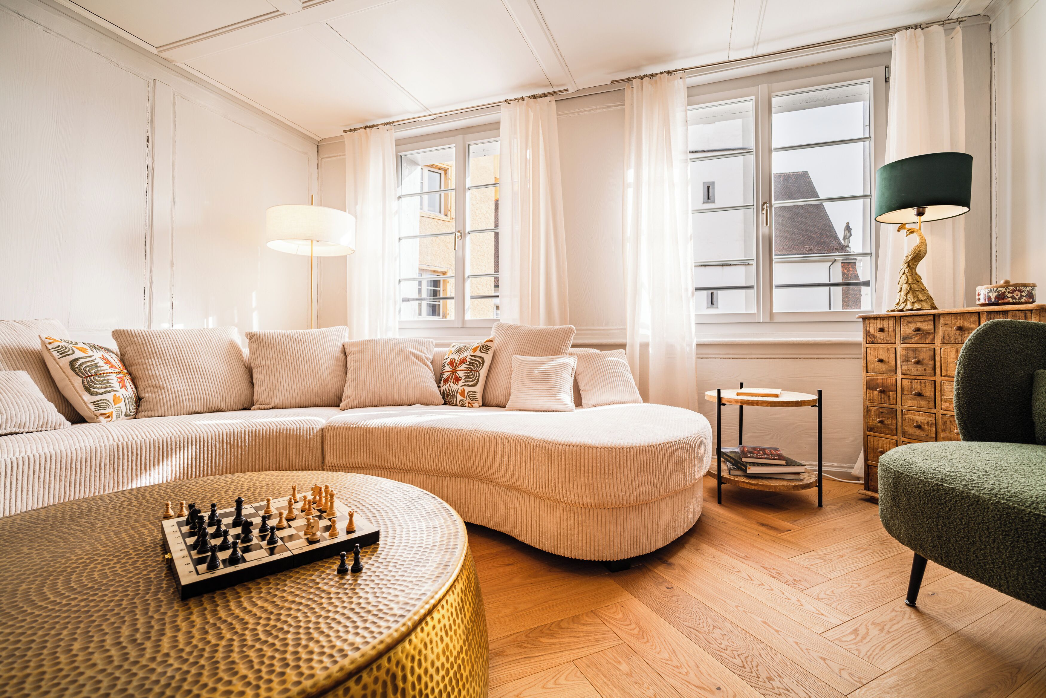 A living room with a sofa, a round coffee table with chess pieces, a lamp, and a view through a window.