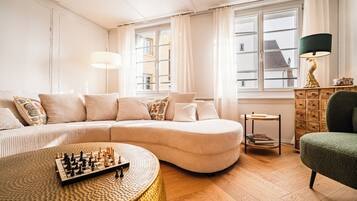 A living room with a sofa, a round coffee table with chess pieces, a lamp, and a view through a window.
