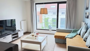 Un salón moderno con una televisión de pantalla plana, una mesa de centro, un sofá y un amplio ventanal con vistas a edificios.