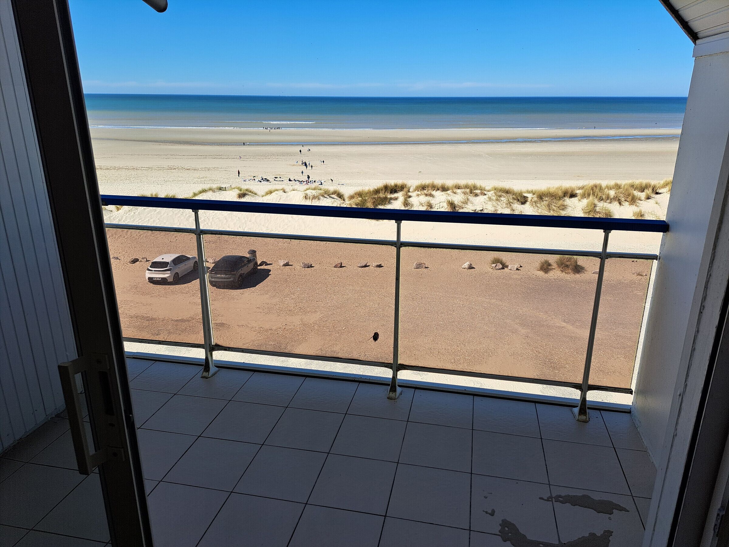 A balcony with a view of a beach, a parked car, and a sandy area with rocks.