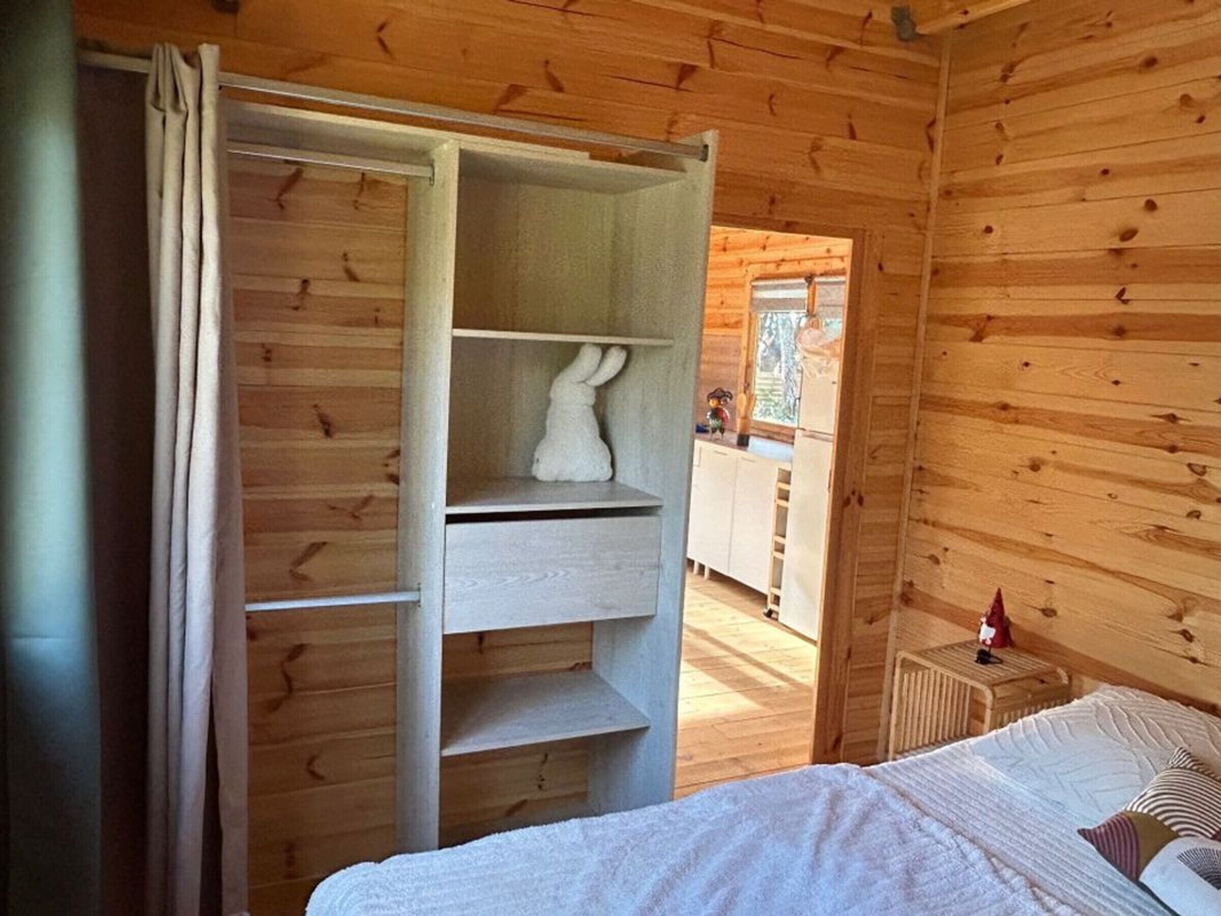 Ein Holzinnenraum mit einem Bett, einem weißen Regal mit einer weißen Hasenfigur und einem durchgehenden offenen Durchgang zur Küche.
