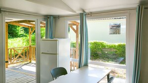 Cocina compacta con refrigerador, una mesa y sillas, una puerta corrediza de vidrio que da a una terraza exterior y vistas a la vegetación.