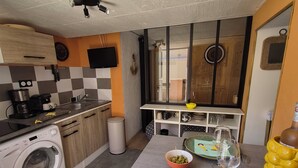 Een compacte keuken met een wasmachine, een spoelbak, een eettafel met een bord en bestek, en een aan de muur bevestigde televisie.