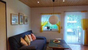 Un salon avec un canapé bleu foncé, une table basse en verre et un tapis à motifs. Il y a une fenêtre avec des rideaux jaunes et une lanterne en papier suspendue.