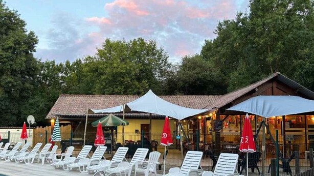 Zona de piscina con sillones de descanso y sombrillas, un edificio de madera con techo de tejas, y un entorno natural de árboles.