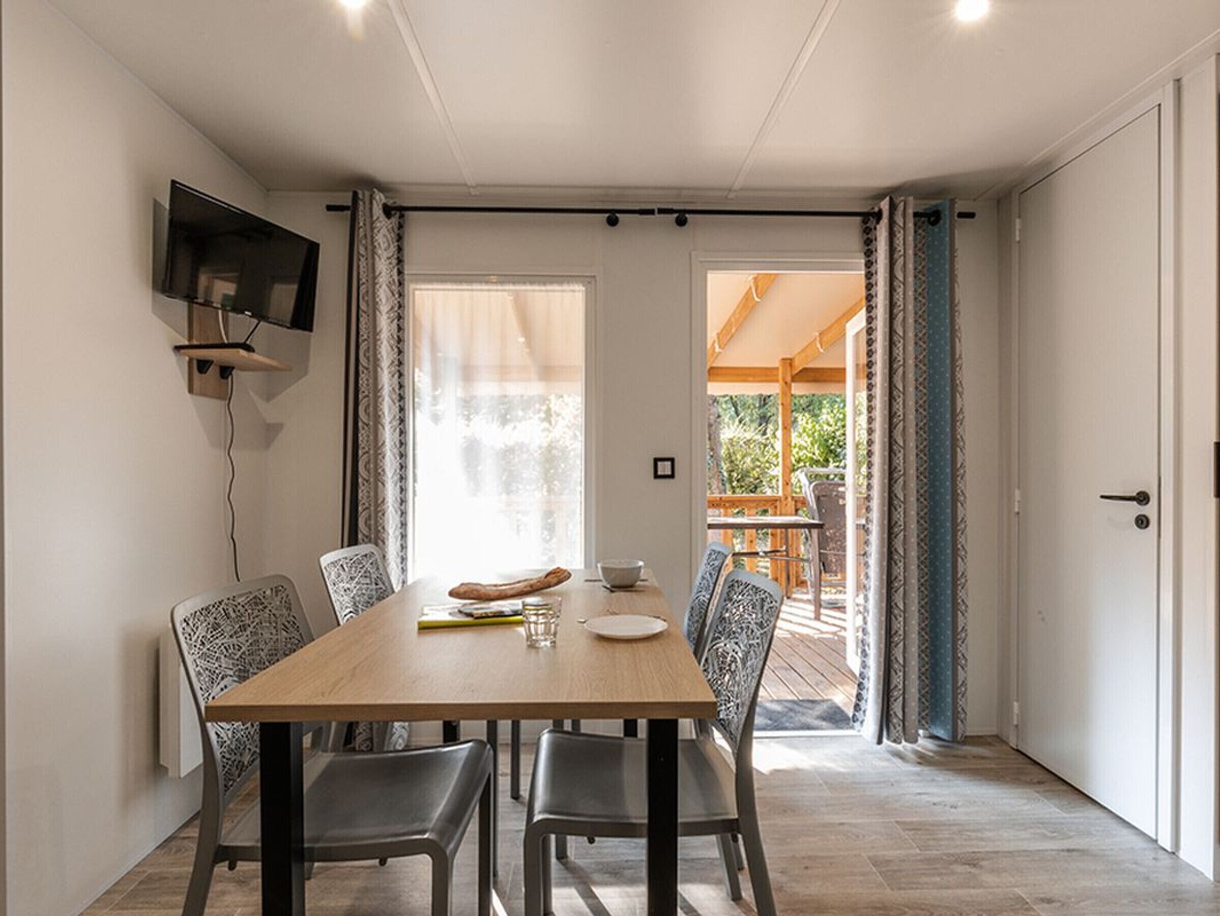 Uma área de refeições com mesa de madeira, quatro cadeiras, uma TV de tela plana na parede e vista para um deck externo.