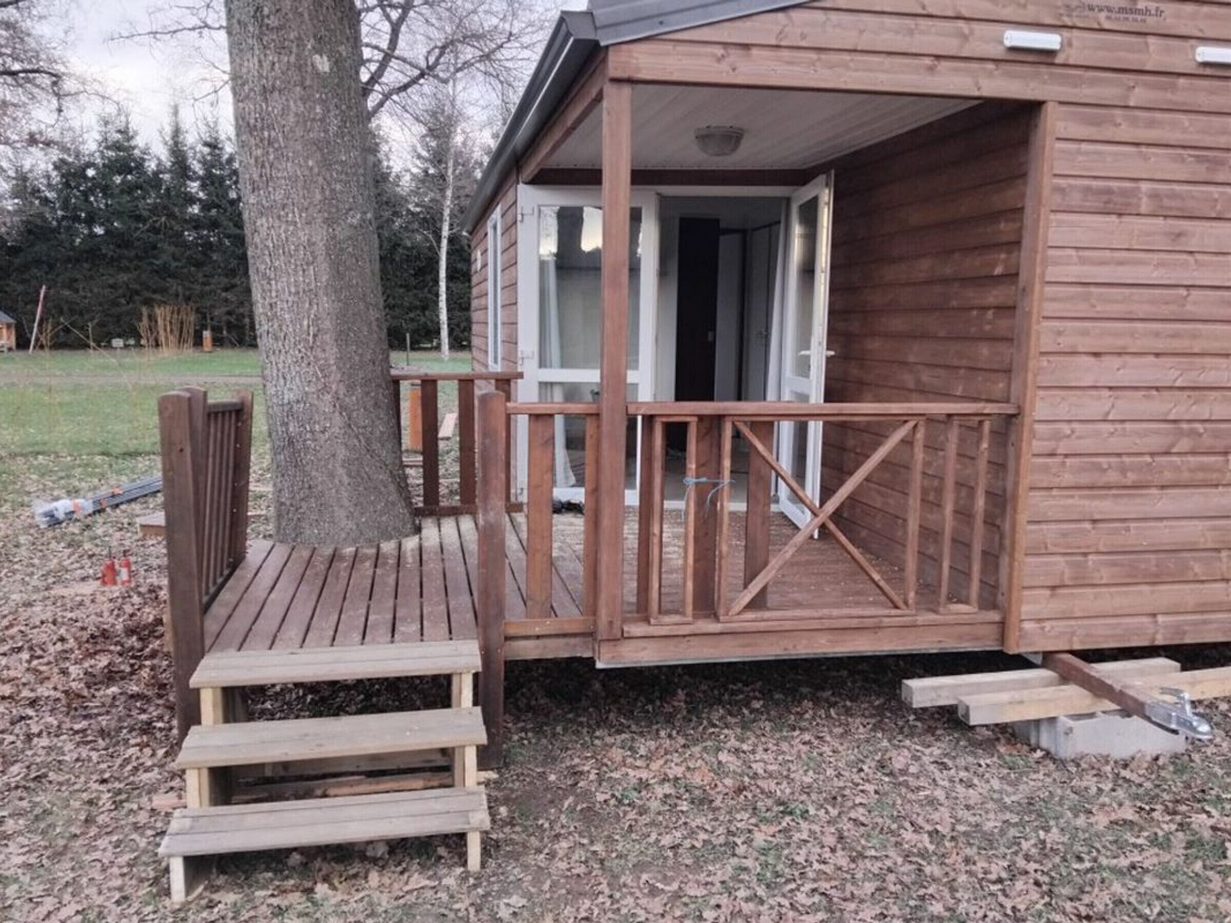 Une cabane en bois avec un porche, un escalier menant à l’entrée et un arbre à proximité.