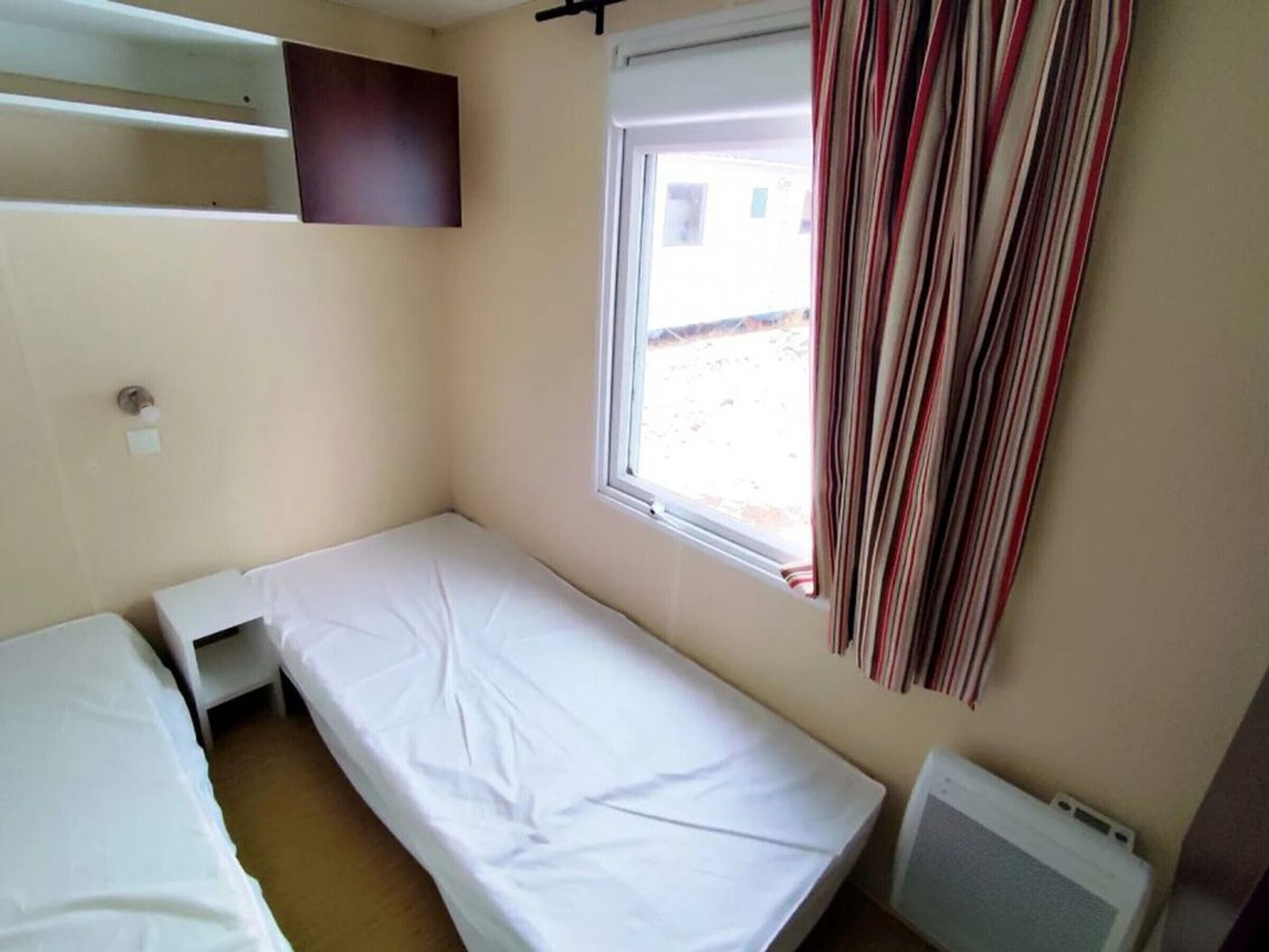 Quarto pequeno com cama de solteiro, janela, radiador e prateleira sobre a cama.
