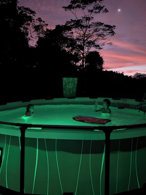 An outdoor hot tub with a green-lit pool, surrounded by trees and a night sky with a crescent moon.