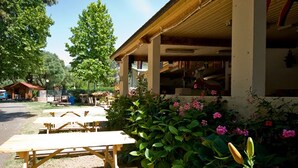 Et overdækket udendørs område med picnicborde, en bygning med saddeltag og en have med blomstrende planter.