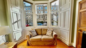 Ein Eckzimmer mit beigefarbenem Sofa, Holzboden und einem Erkerfenster mit Blick auf die Stadt.