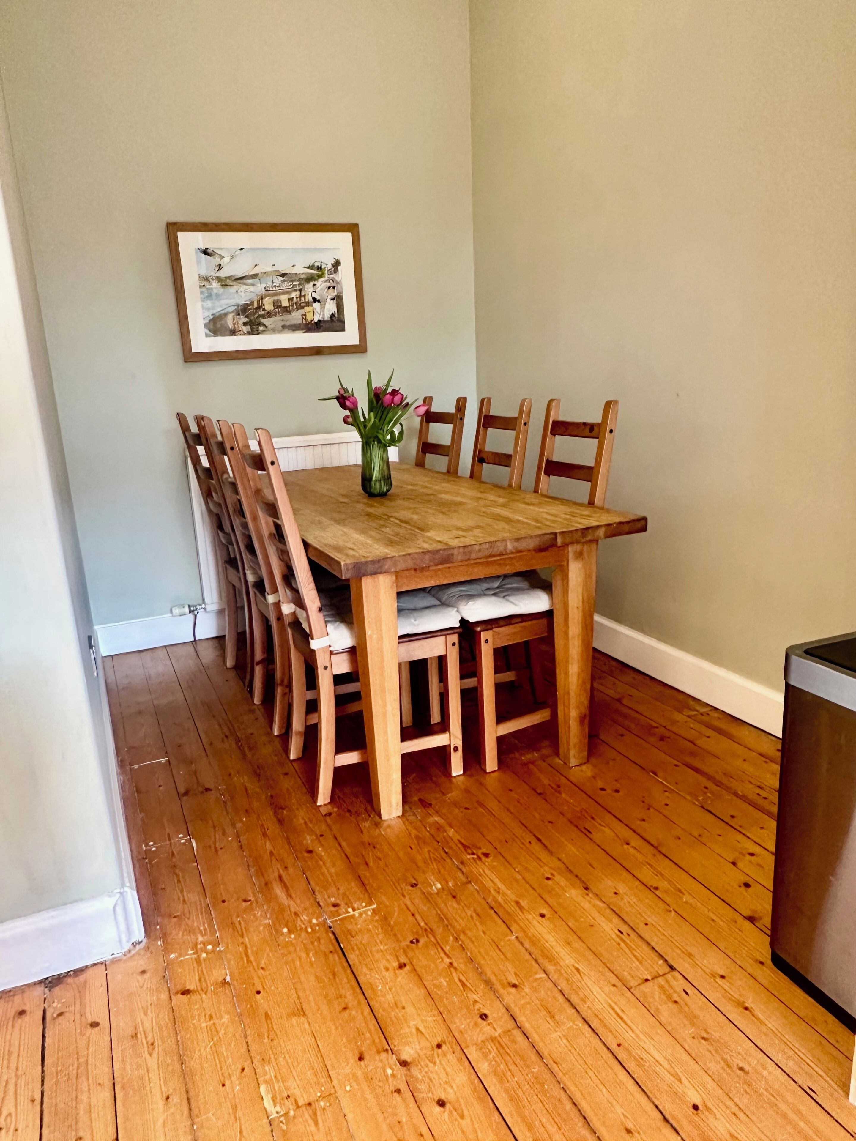 A dining area with a wooden table and chairs, a vase with flowers, and a framed picture on the wall.