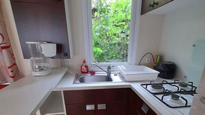 Pieni, käytännöllinen keittiö, jossa on pesuallas, liesi ja ikkuna, josta avautuu näkymä vehreään maisemaan.