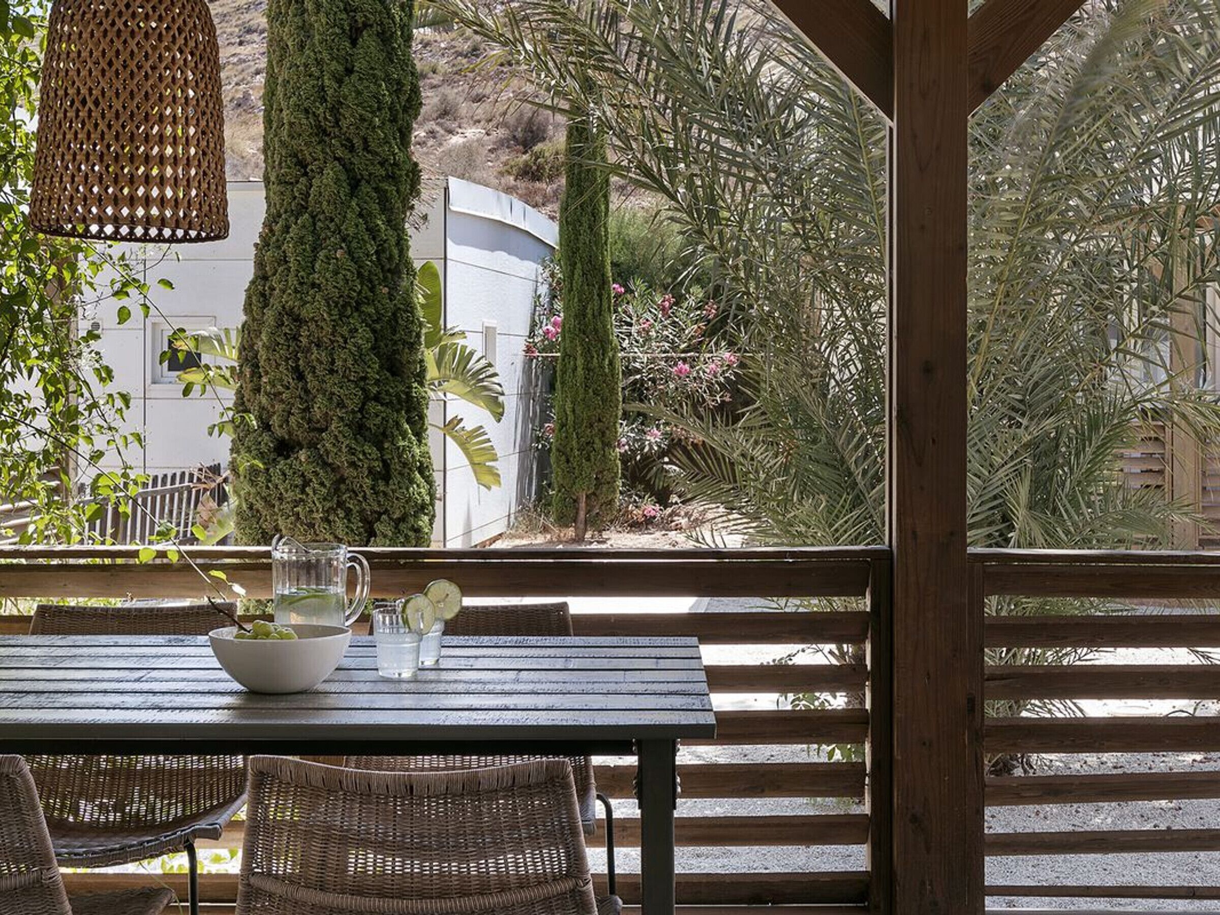 A wooden table with wicker chairs, a hanging light fixture, and a view of a garden with various plants and a tall tree.