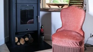 A room with a black wood-burning stove, a wicker chair, and a window with a view of trees. - Mobile home, pets welcome - 5 guests (La Châtre)