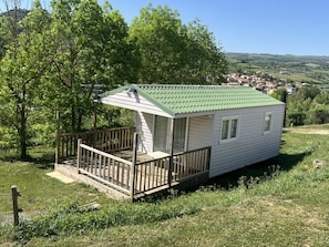A small, single-story cabin with a green roof, a wooden deck, and a view of a rural landscape.