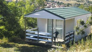 Uma cabana pequena, térrea, com telhado inclinado, varanda com guarda-corpo e vista para uma paisagem rural.