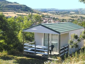 A small, single-story cabin with a sloped roof, a porch with a railing, and a view of a rural landscape. - Mobile home, pets welcome - 4 guests (Saint-Georges-de-Luzençon)