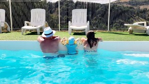 Piscina cercada, cadeiras de descanso brancas e um monte gramado ao fundo.