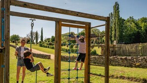 Una estructura de juegos de madera en un espacio al aire libre con césped, una pared de piedra y árboles al fondo.