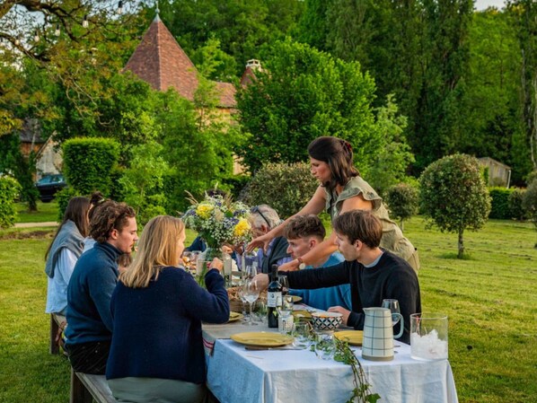 A group of people seated at an outdoor table with a white tablecloth, surrounded by greenery and a building with a conical roof in the background.