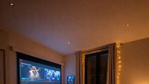 Un dormitorio con una televisión grande montada en la pared, una cama con una manta, una mesita de noche con una lámpara y una ventana con cortinas.