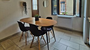 Un comedor con una mesa redonda, cuatro sillas, una planta en maceta, un espejo y una ventana que ofrece vistas a un edificio de ladrillo.