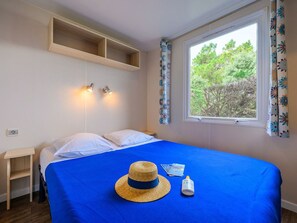 A bedroom with a bed, a window with curtains, a bedside table, and a wall-mounted shelf. - Mobile home - pets allowed - 4 guests (Saint-Jean-de-Monts)