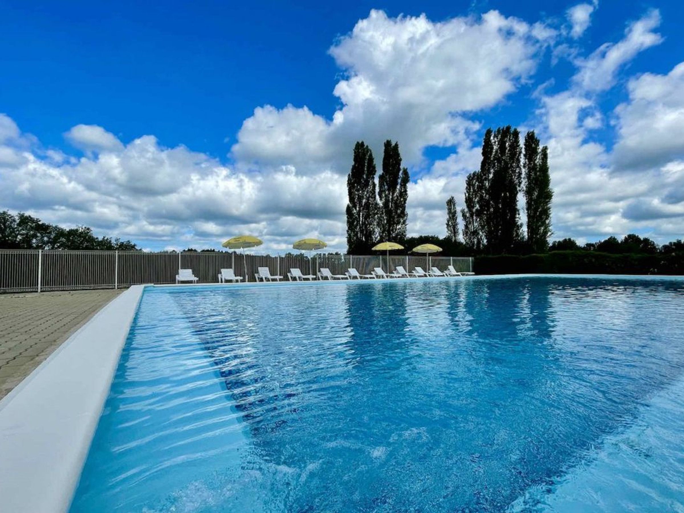 Una piscina con sillones de descanso y sombrillas, rodeada por una valla y árboles bajo un cielo parcialmente nublado.