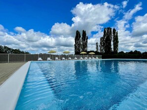 Ein Pool mit Liegestühlen und Sonnenschirmen, umgeben von einer Hecke und Bäumen unter einem bewölkten Himmel.