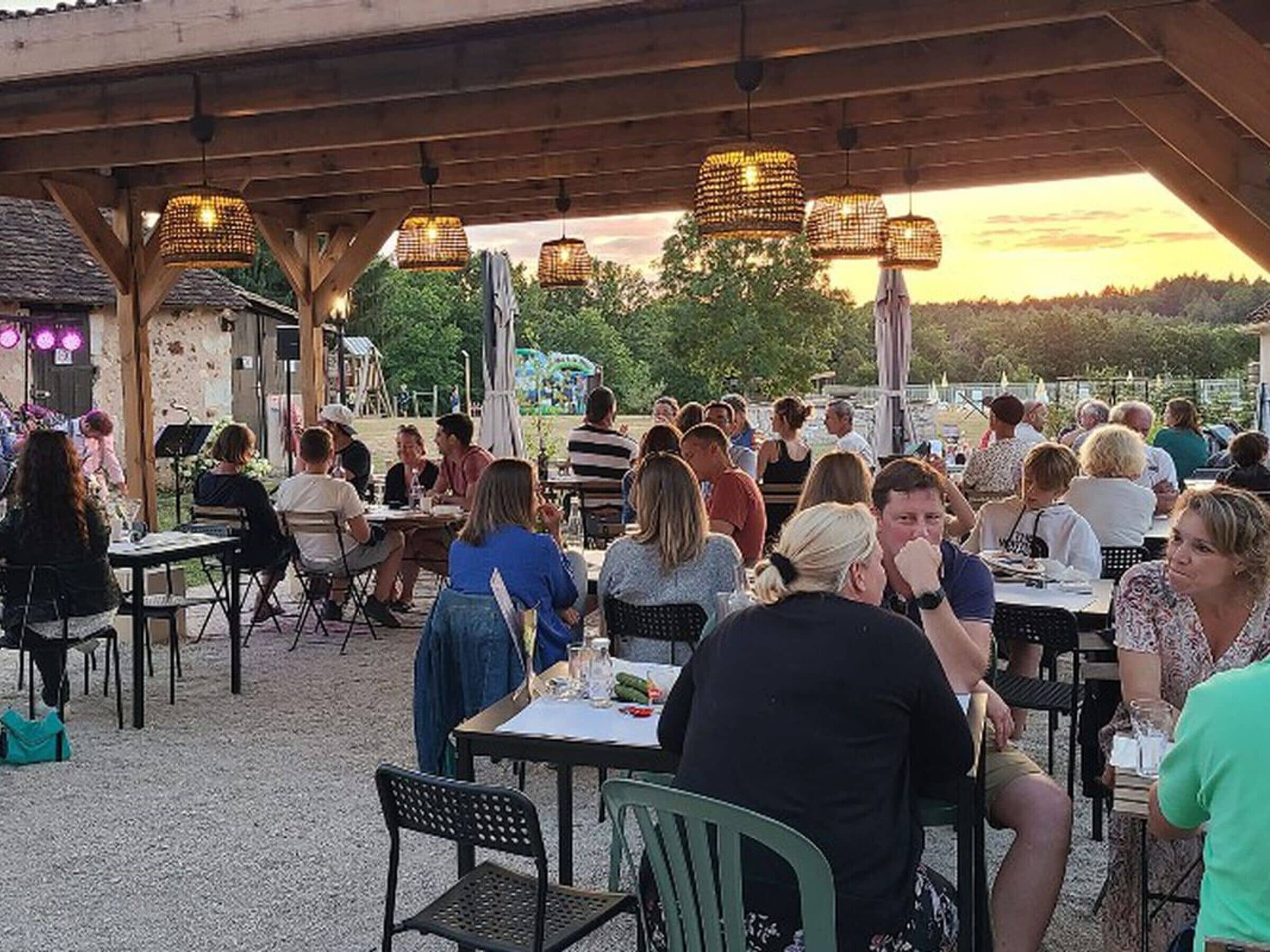 An outdoor dining area with tables and chairs, covered by a wooden structure with hanging lights.