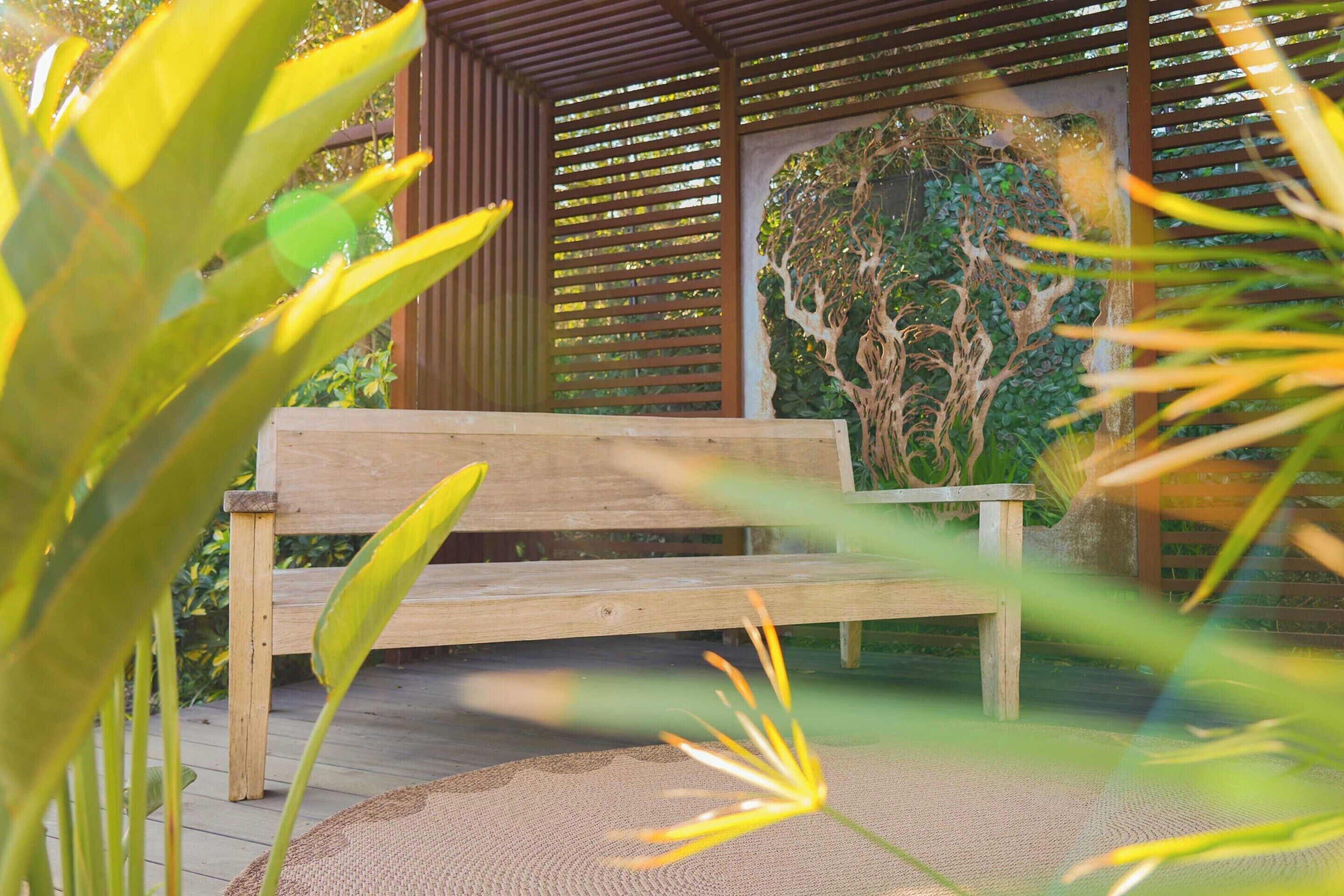 A wooden bench in a covered outdoor area with a wooden slatted roof and a wall with a green leafy design.