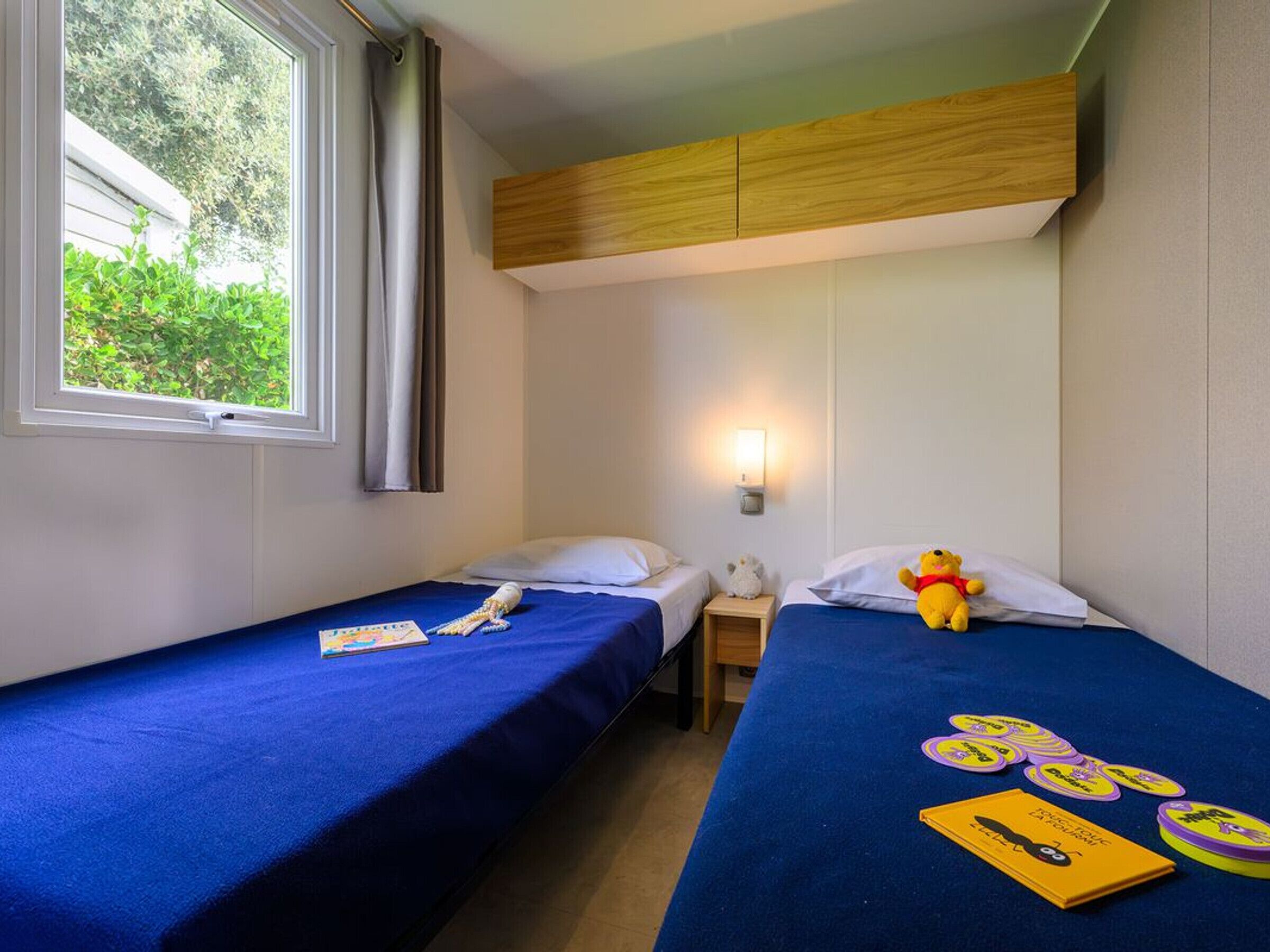A bedroom with two beds, a window with curtains, a wall-mounted light, and a stuffed toy on one bed.