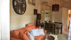 Interior de madera con un gran reloj de pared, sofá naranja, mesa de comedor y vigas de madera en el techo.