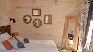 Dormitorio de una cabaña de madera con una cama, almohadas, un espejo fijado a la pared y una ventana con cortinas.