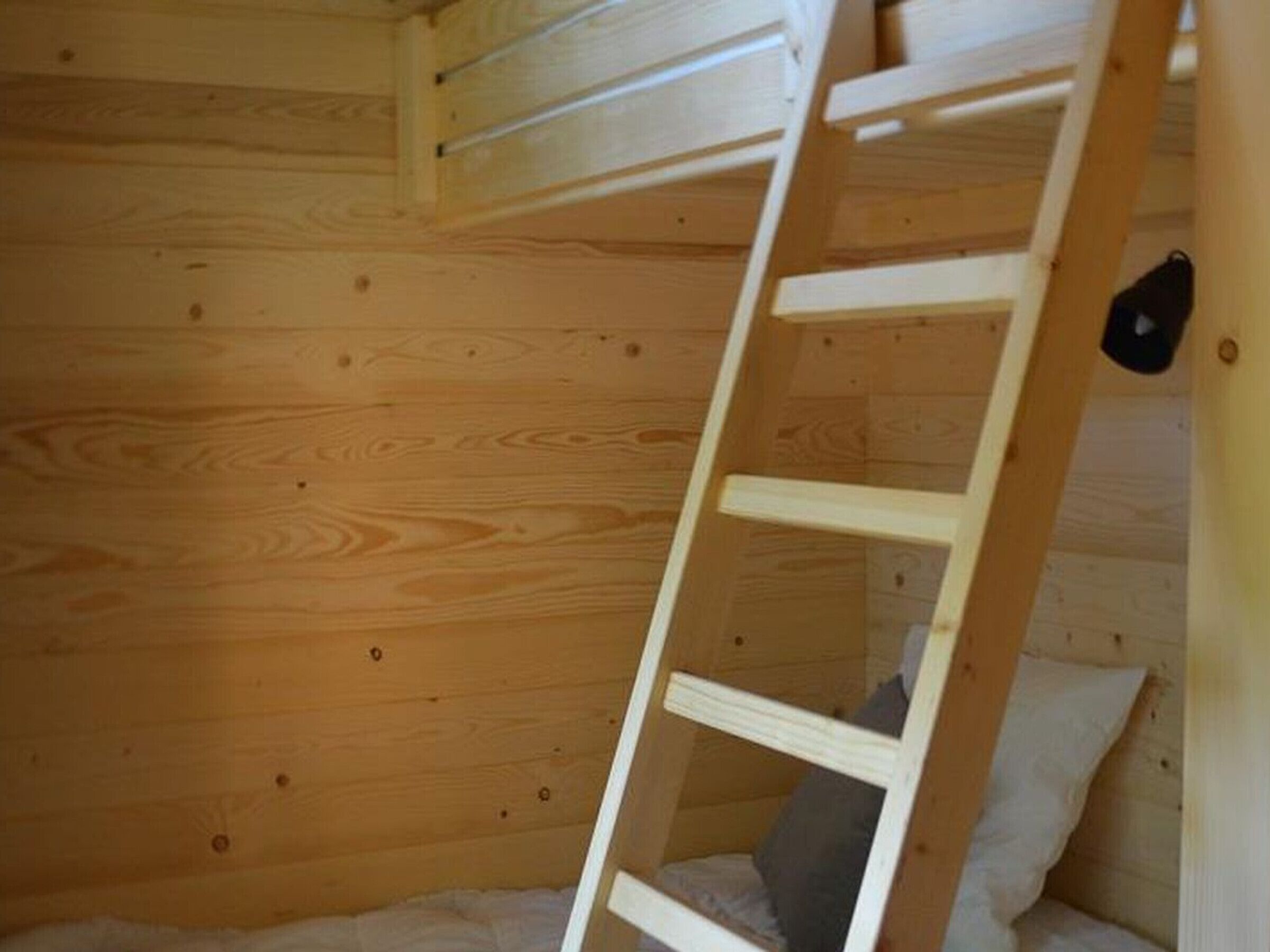 A wooden bunk bed room with a ladder, pillows, and a small black object on the wall.