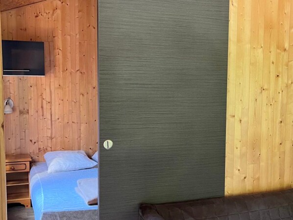A wooden cabin interior with a bed, a TV mounted on the wall, and a door with a round handle.