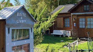 Uma cabana de madeira com uma pequena biblioteca identificada como 'BOITE A LIVRES' e um banco no quintal.