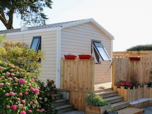 A single-story mobile home with a front porch, a wooden fence, and a small garden area with flowers. - Cozy mobile home with TV - 4 guests (Sarzeau)