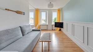 Ett vardagsrum med en grå soffa, ett litet träbord, en tv och stora fönster med gardiner.