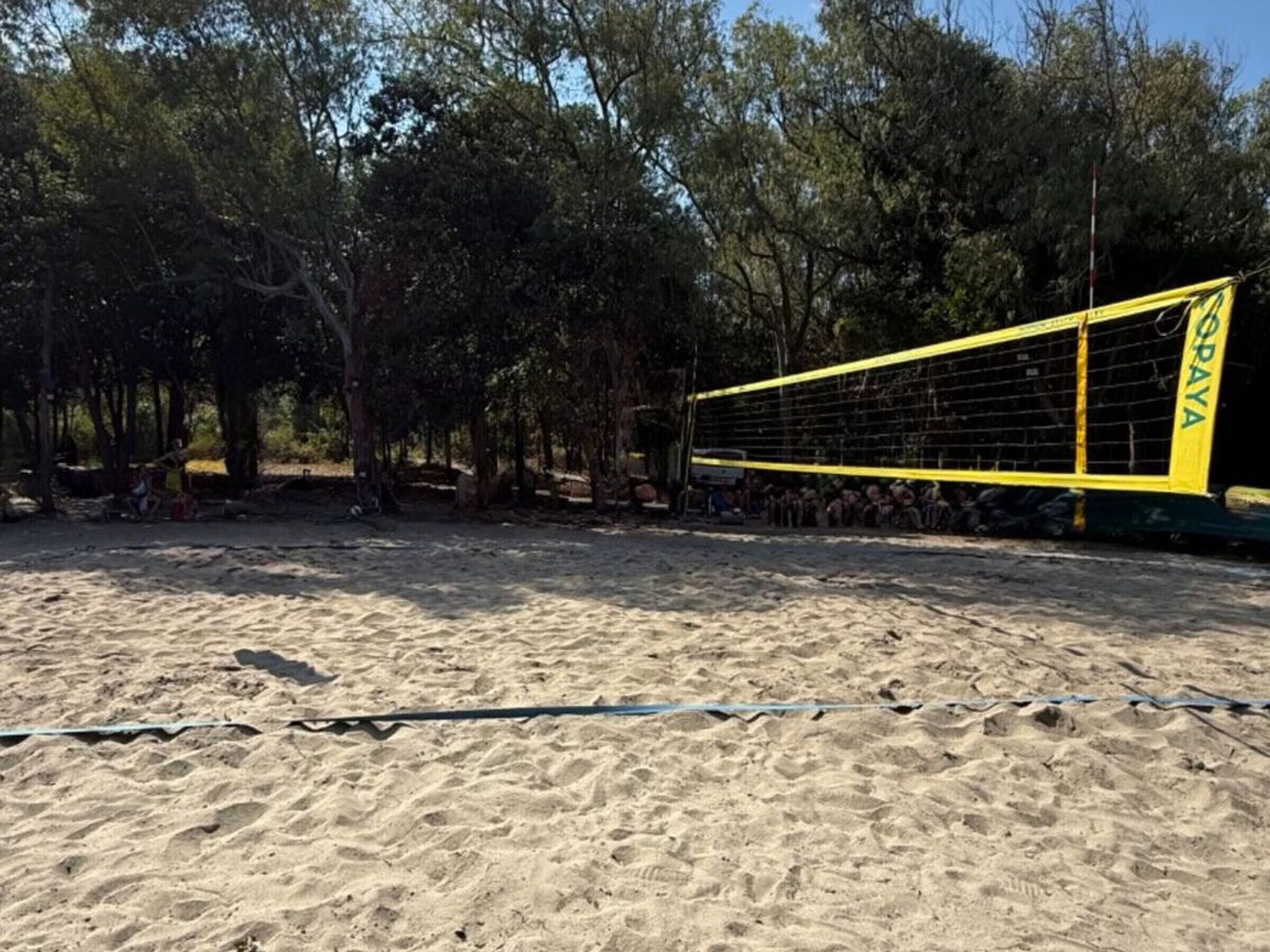 Un terrain de beach-volley sablonneux avec un filet portant l’inscription « CORAYA » et des arbres en arrière-plan.